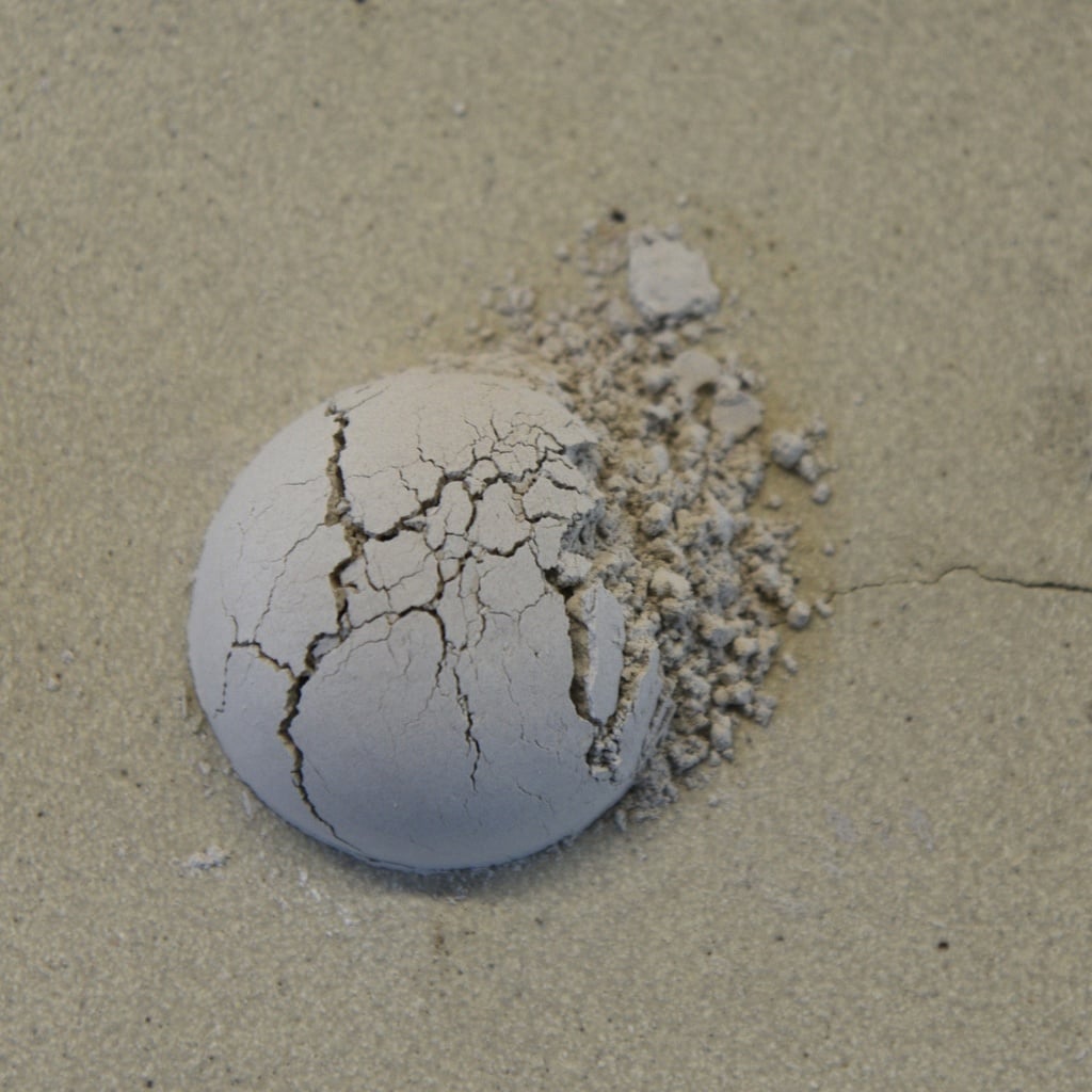 Cracked ball of clay on a sandy surface, showing texture and natural imperfections, ideal for sculpting or casting projects.