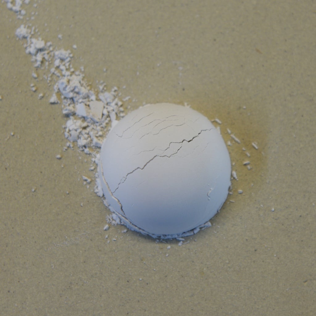 Cracked white clay sphere on a sandy surface, surrounded by small clay debris, showcasing ceramic imperfections.