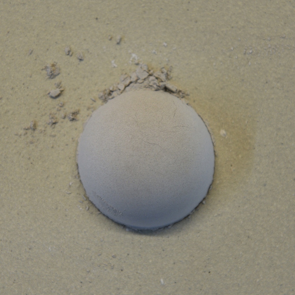 A smooth, rounded ball of clay surrounded by fine particles, resting on a light, sandy surface, ideal for sculpting.