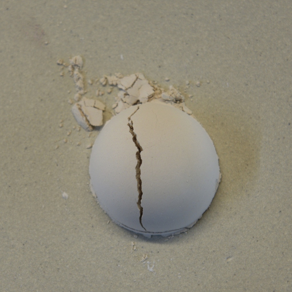 Cracked ceramic sphere resting on a light surface, surrounded by small pieces of clay, showcasing pottery damage.